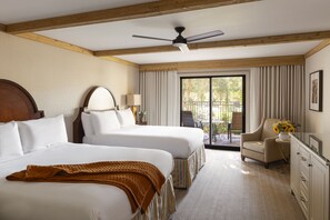Une chambre d’hôtel avec deux lits, un ventilateur de plafond, une grande fenêtre avec des rideaux, une chaise et une petite table sur laquelle est posé un vase de fleurs.