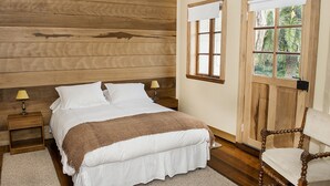 Un dormitorio con cabecera de madera, cama con ropa blanca, mesita de noche con lámpara y ventana con vista a vegetación.