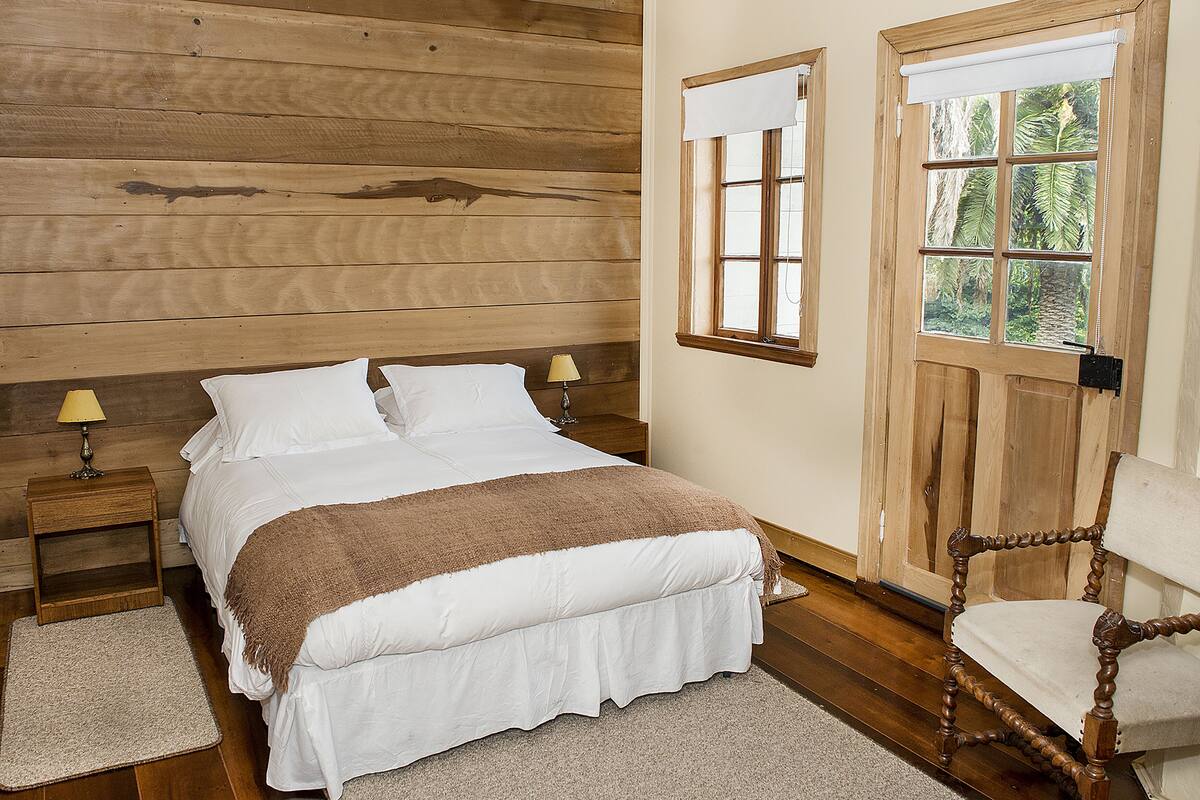 Un dormitorio con cabecera de madera, cama con ropa blanca, mesita de noche con lámpara y ventana con vista a vegetación.