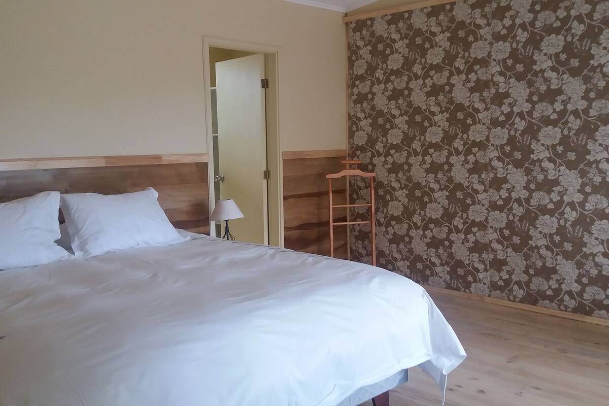 Un dormitorio con una cama grande, un cabecero de madera, una mesita de noche con lámpara y un papel tapiz con estampado floral.