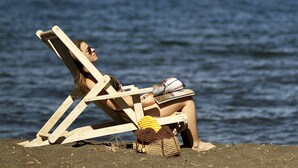 Escena de playa con dos personas relajándose en sillas de madera, un bolso de playa y un sombrero de paja en la arena, con una montaña y el océano al fondo.