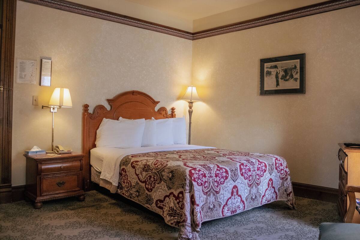 A hotel room with a wooden bed, two bedside lamps, a nightstand, a dresser, and a framed picture on the wall.