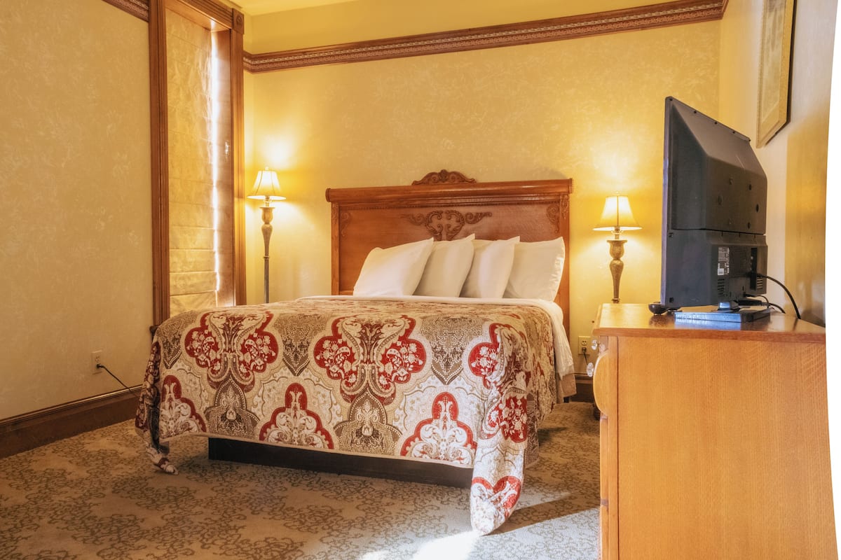 A hotel room with a bed, two bedside lamps, a television on a wooden dresser, and a carpeted floor.