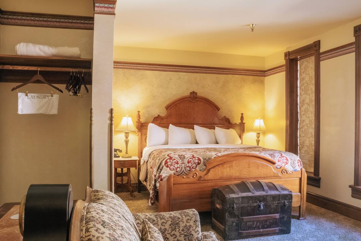 A bedroom with a wooden bed, two bedside tables with lamps, a hanging laundry bag, and a vintage suitcase.