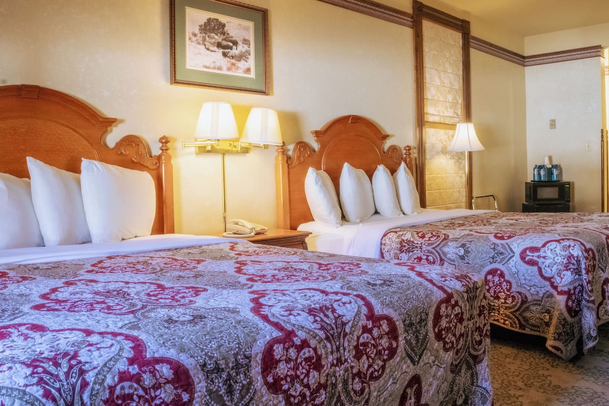 A hotel room with two beds, each with a wooden headboard, white bedding, and a patterned bedspread. There are bedside lamps, a telephone, and a small table with a microwave and beverages.