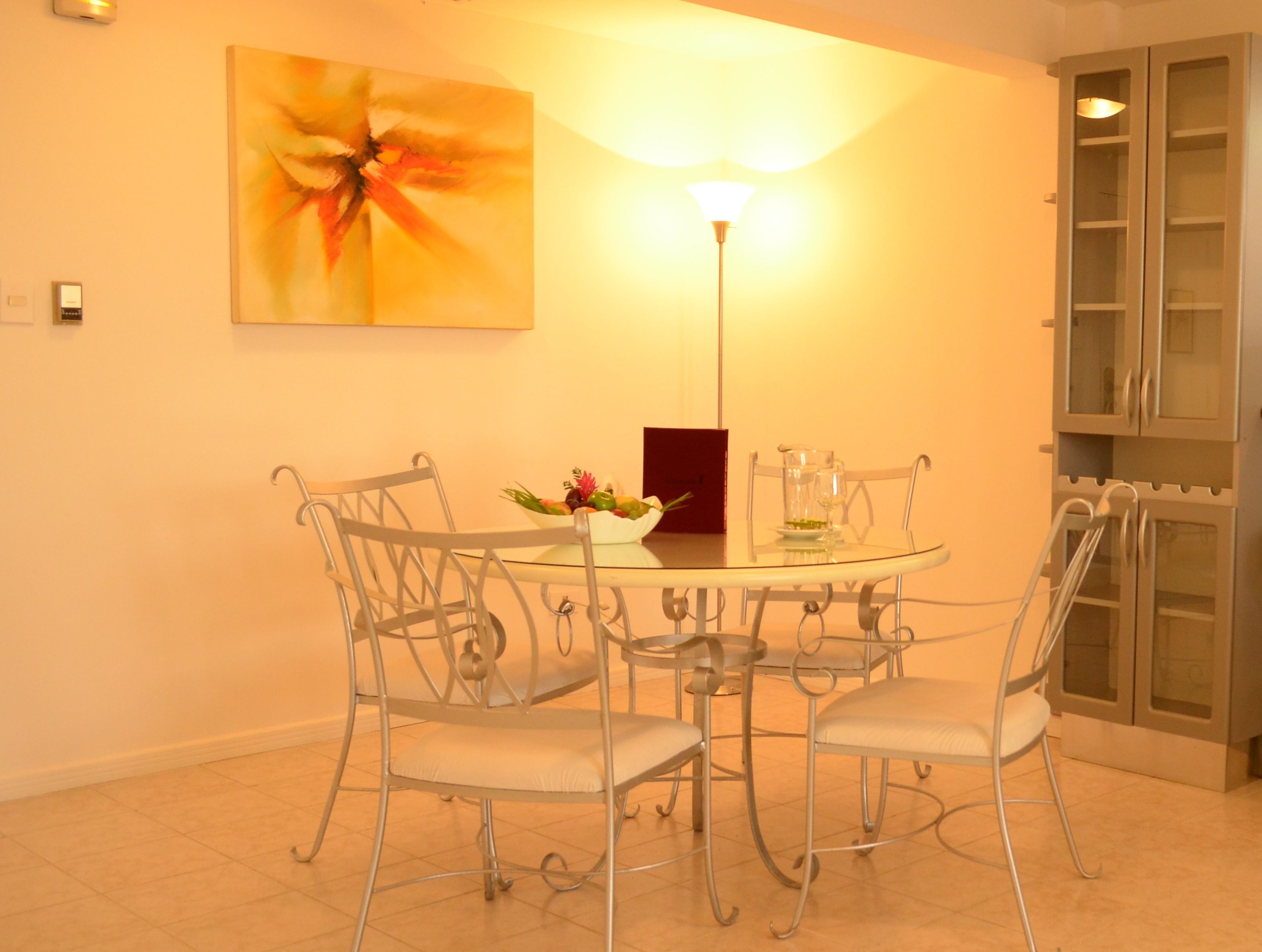 A dining area with a round glass table, four metal chairs, a flower centerpiece, and a wall-mounted painting.