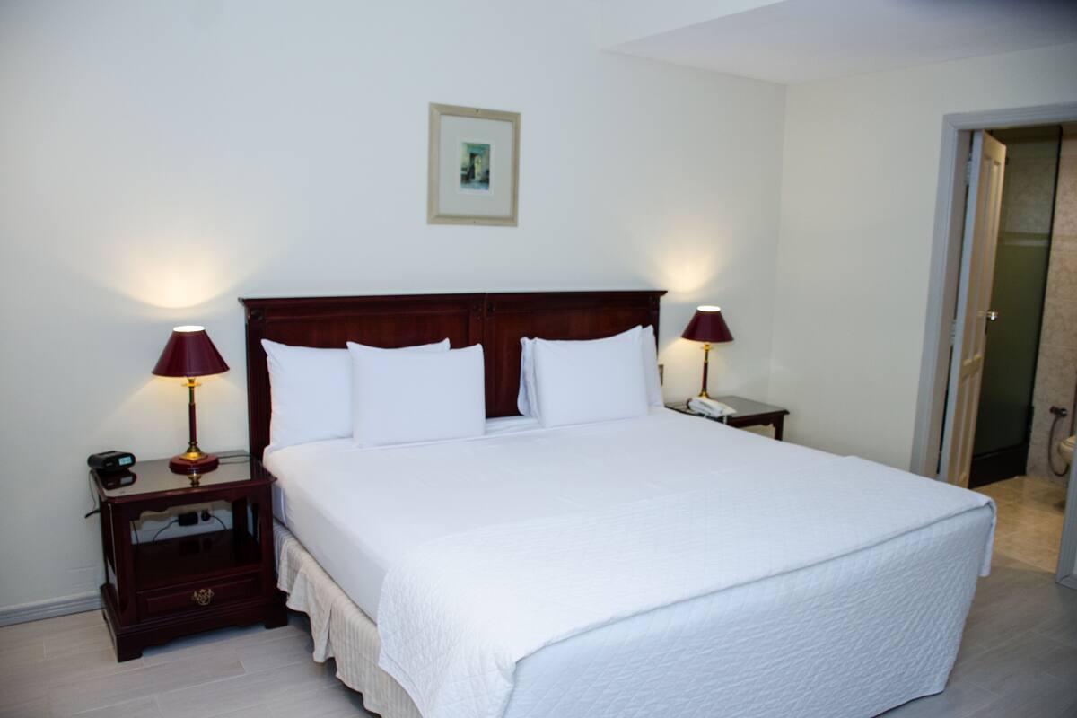 Una cama grande con ropa blanca, dos mesitas de noche con lámparas y un cuadro enmarcado en la pared.