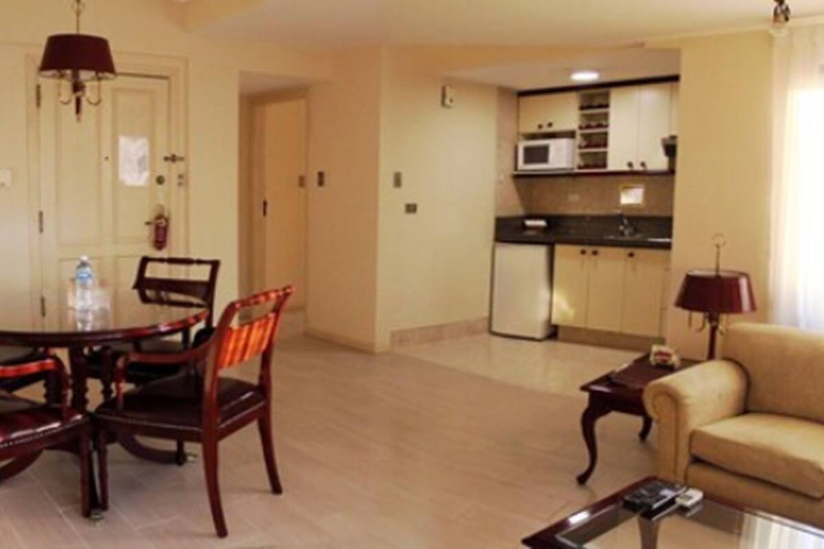Una sala de estar con una mesa de comedor y sillas, un sofá, y una cocina al fondo, además de una lámpara.