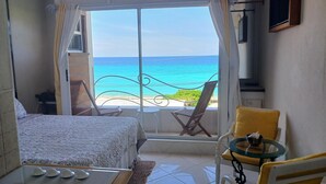 A room with a bed, a small table with decorative items, a chair, and a view of the ocean through a large window. - Coral Lane Beach Apartments (Paynes Bay)