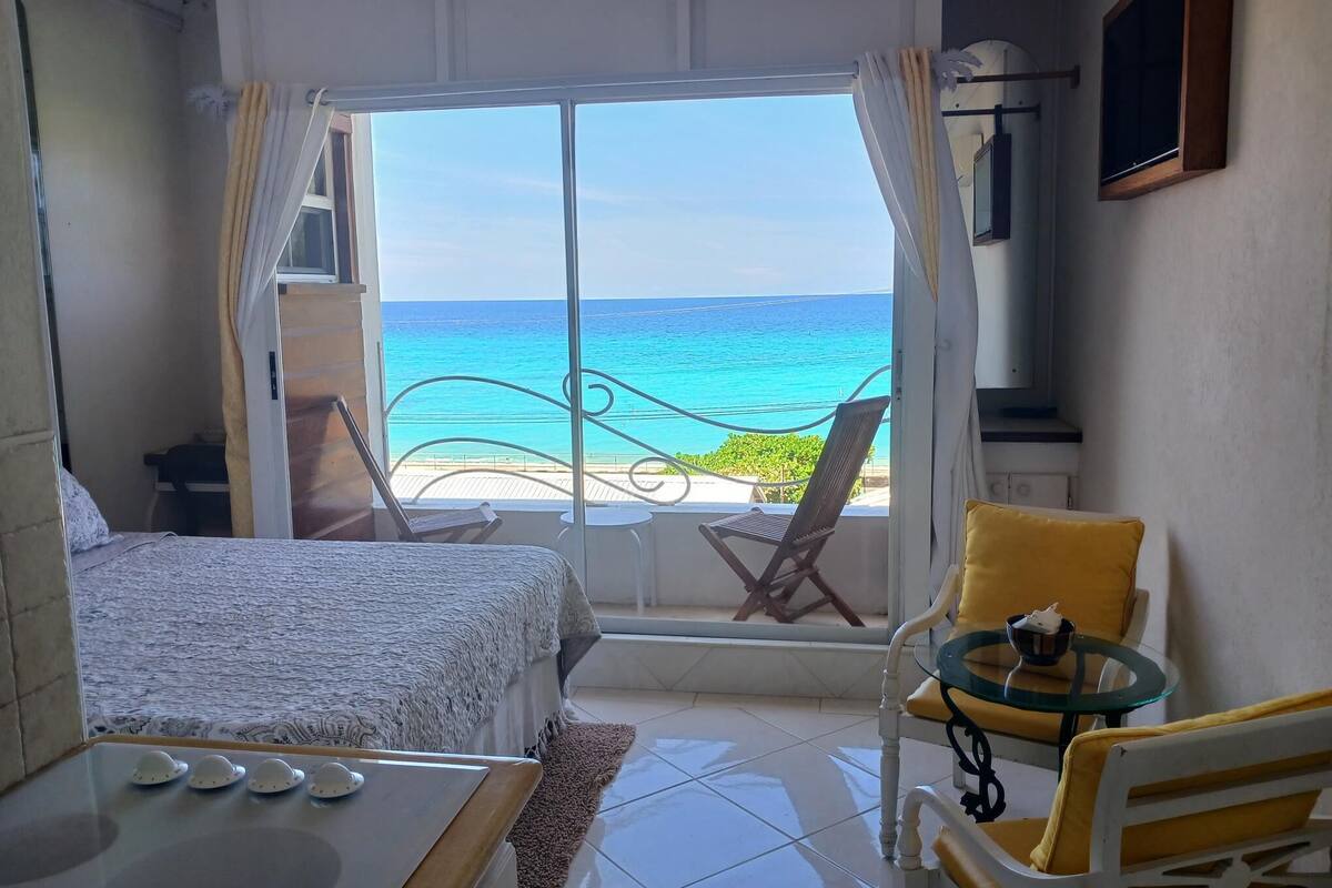 A room with a bed, a small table with decorative items, a chair, and a view of the ocean through a large window.
