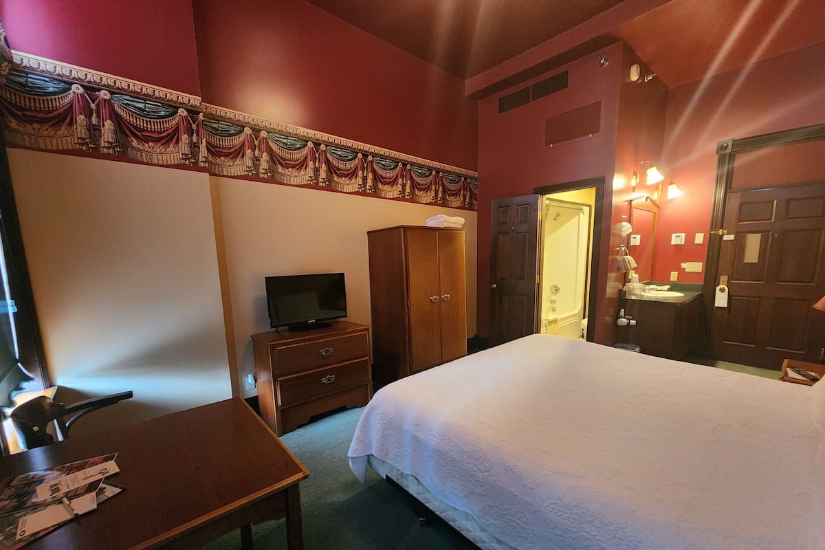 A hotel room with a bed, a TV on a wooden dresser, a wardrobe, and a bathroom visible through an open door.