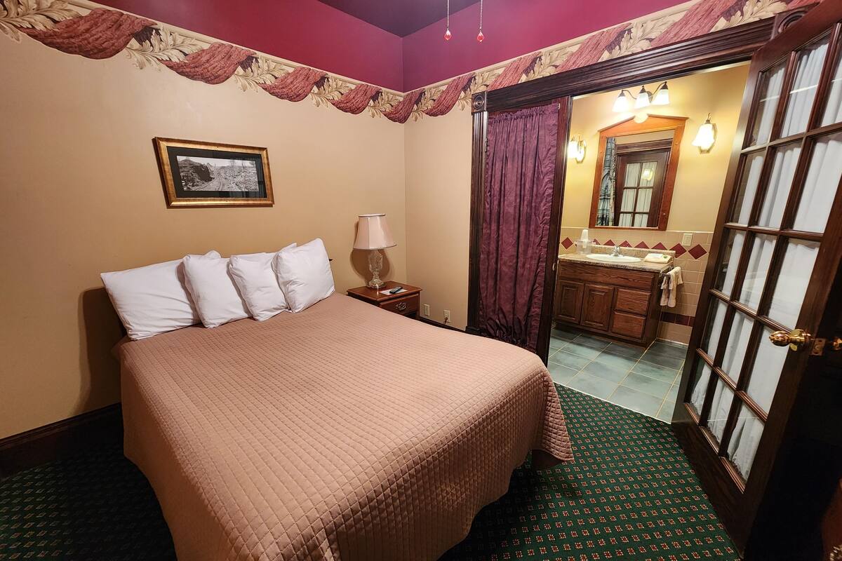 A hotel room with a bed, a nightstand, a lamp, a mirror, and a bathroom visible through an open door.