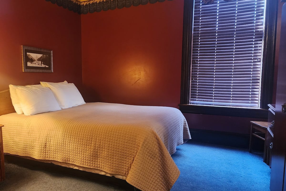 A bedroom with a bed, a nightstand, a lamp, a picture on the wall, and a window with blinds.