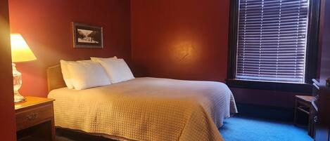 Un dormitorio con cama, mesita de noche, lámpara, un cuadro en la pared y una ventana con persianas.