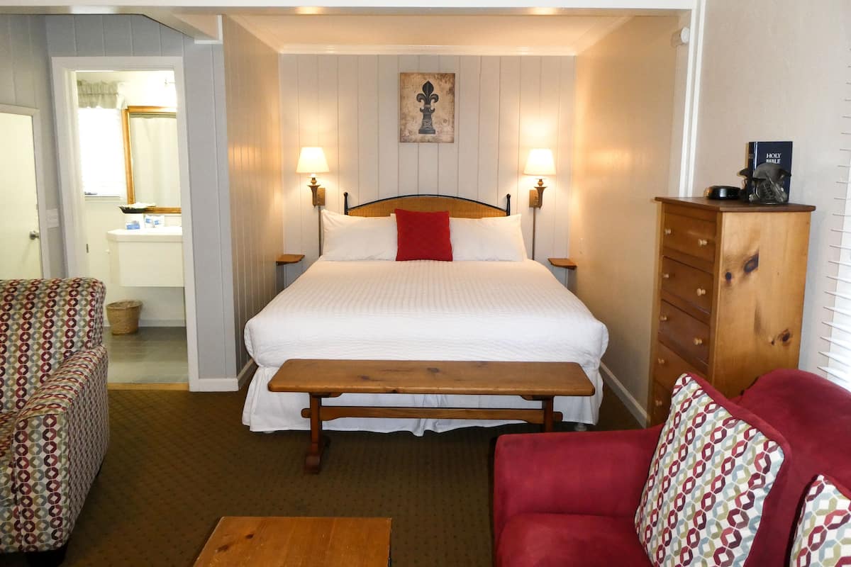 A hotel room with a large bed, two bedside lamps, a wooden dresser, a red chair, and a small table.