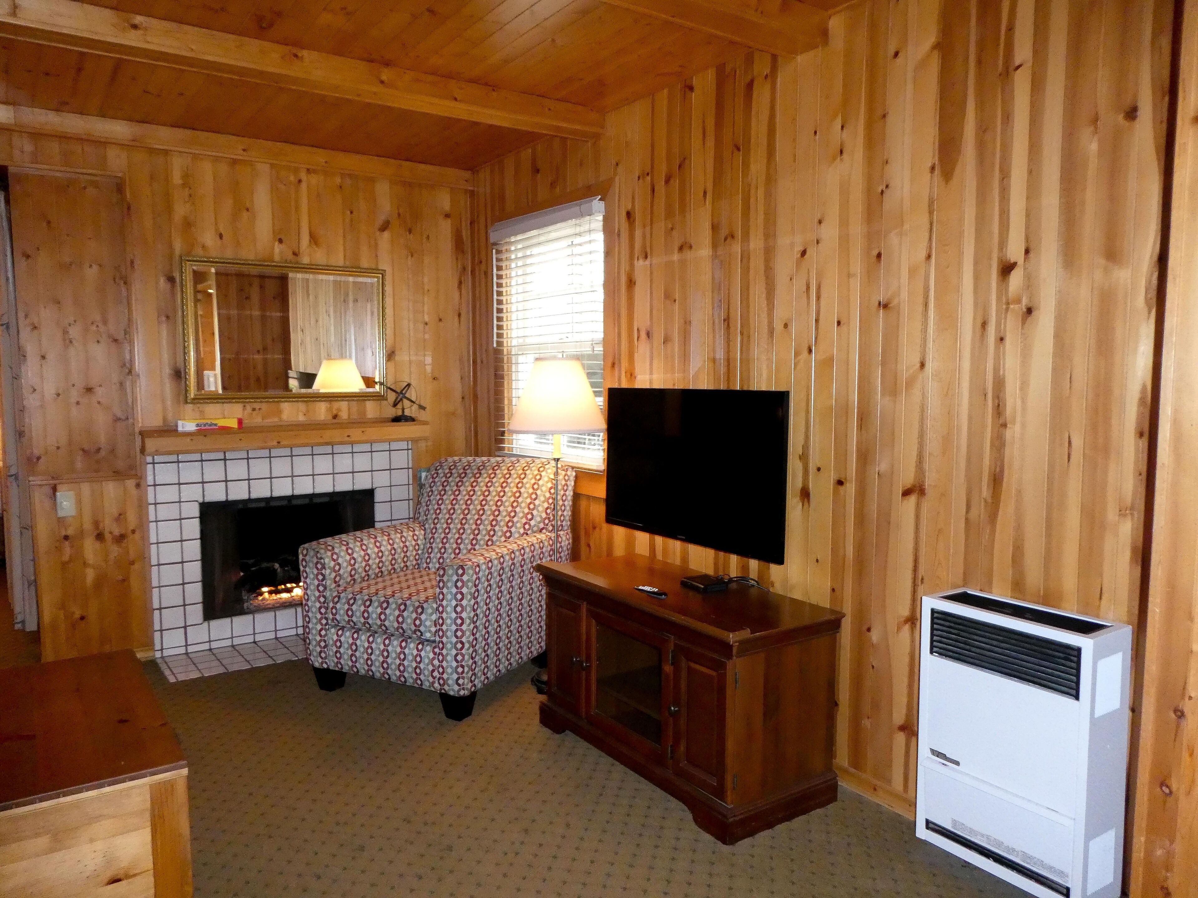 A cozy room with wooden walls, a tiled fireplace, a patterned armchair, a television, and a white air conditioning unit.
