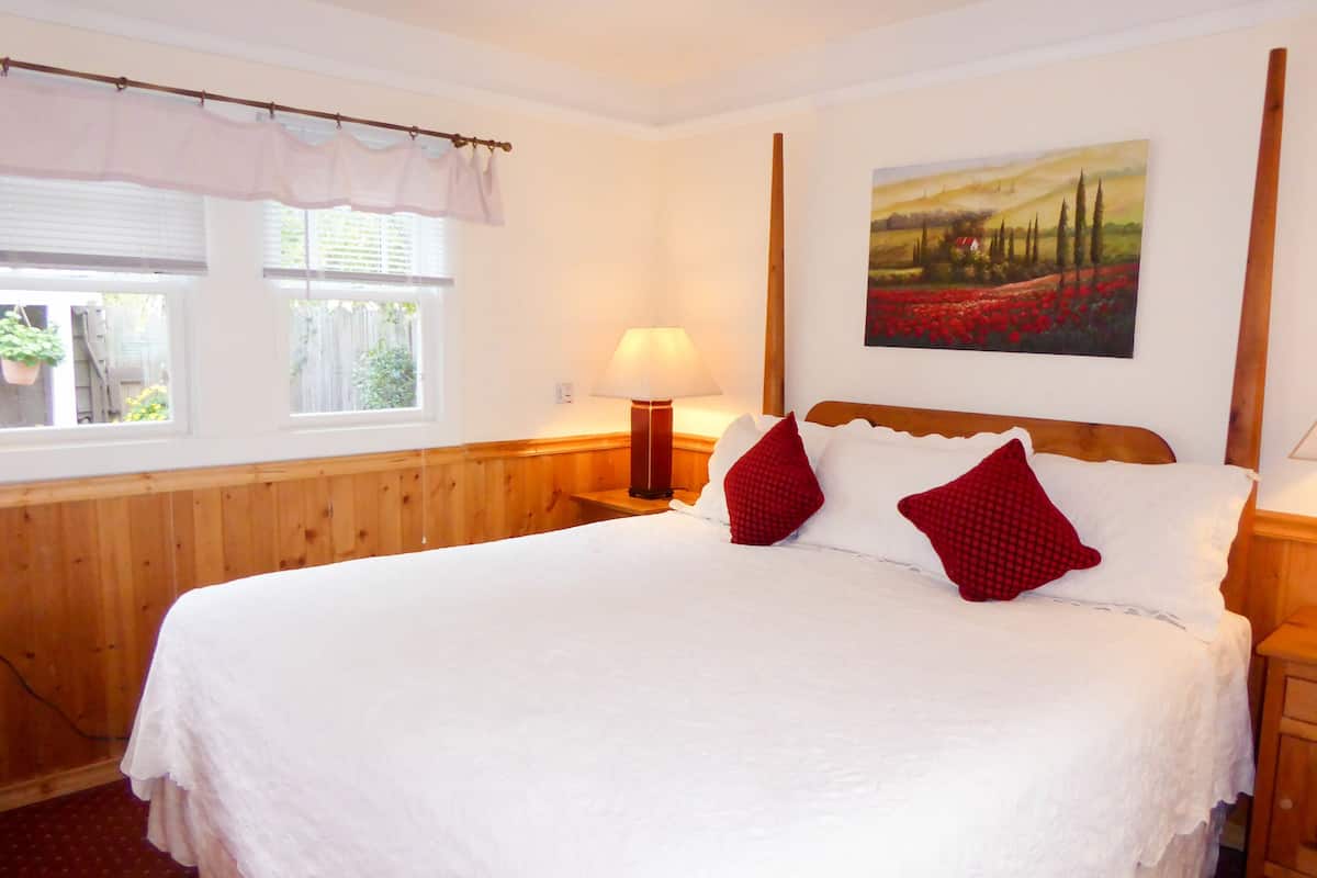 A neatly made bed with white linens and red pillows, a bedside lamp, and a painting of a landscape on the wall.