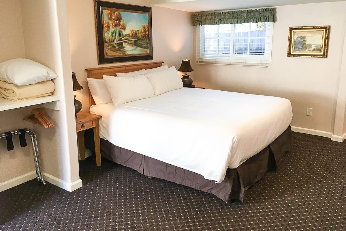 A neatly made bed with white linens, a wooden headboard, and a nightstand with a lamp. A painting hangs above the bed, and a window with curtains is visible.