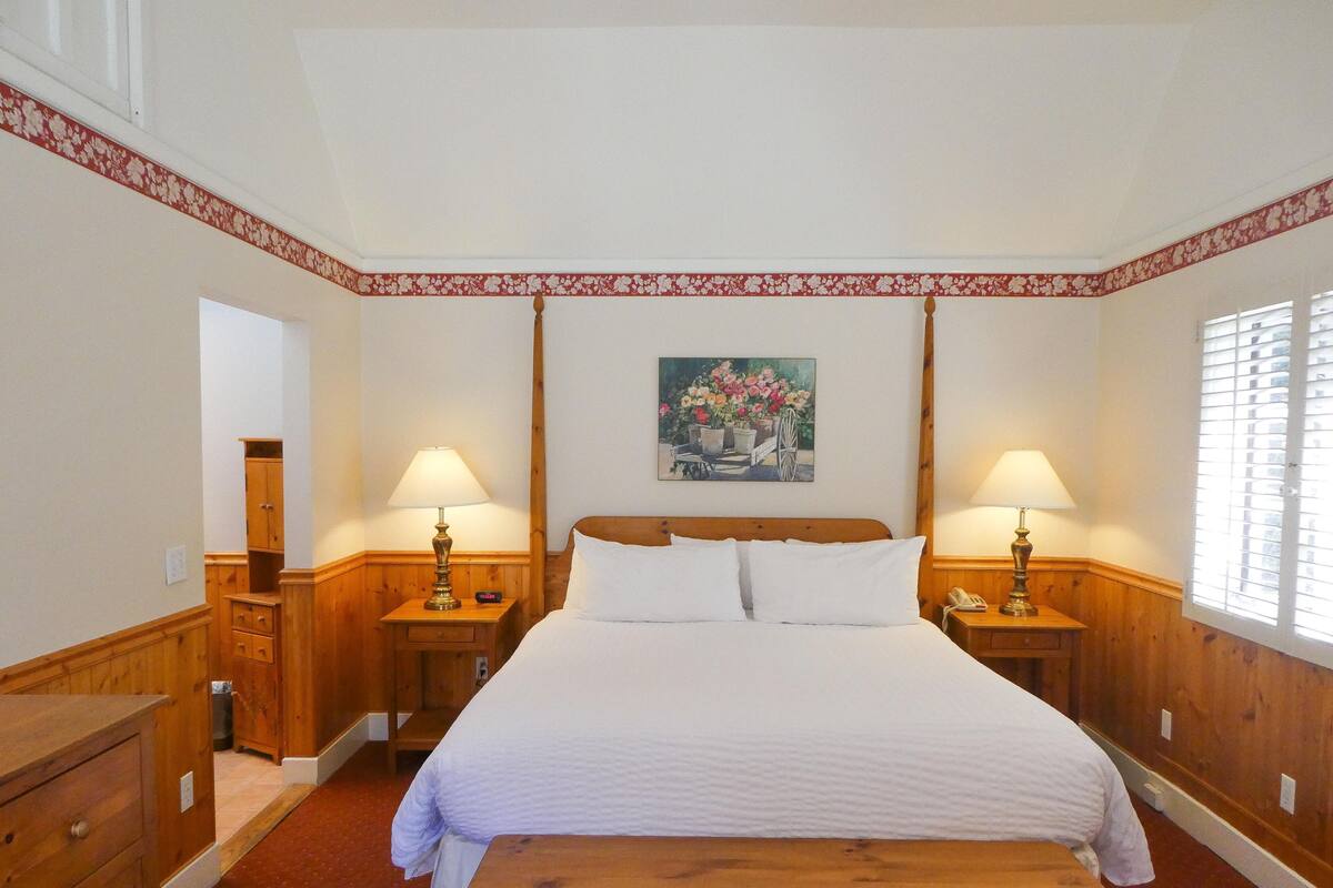 A hotel room with a large bed, two bedside tables with lamps, a wooden bench, a wooden dresser, and a painting on the wall.