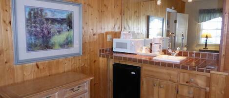 A wooden-paneled kitchen with a microwave, coffee maker, and a sink. There is a framed painting on the wall and a wooden cabinet with drawers.