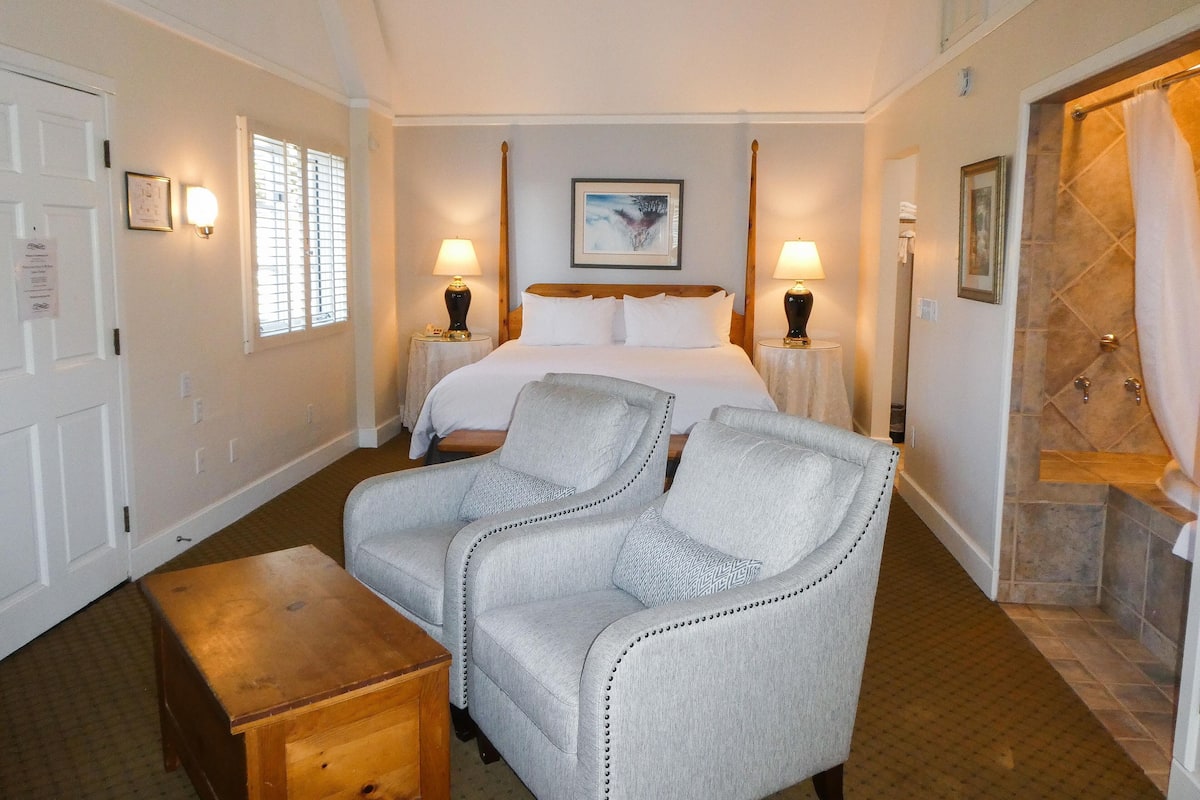 A hotel room with a bed, two armchairs, a wooden side table, two bedside lamps, a painting on the wall, a shower area, and a door with a sign.