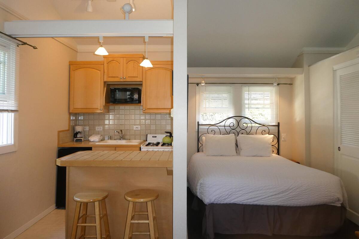 A compact kitchen with wooden cabinets, a microwave, and a gas stove. There are two stools and a small bar area. The adjacent room features a bed with white bedding and a metal headboard.