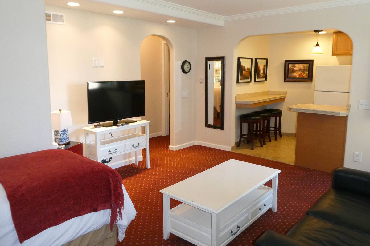 A hotel room with a bed, a TV, a small white table, a red carpet, a kitchenette, and a dining area with stools.