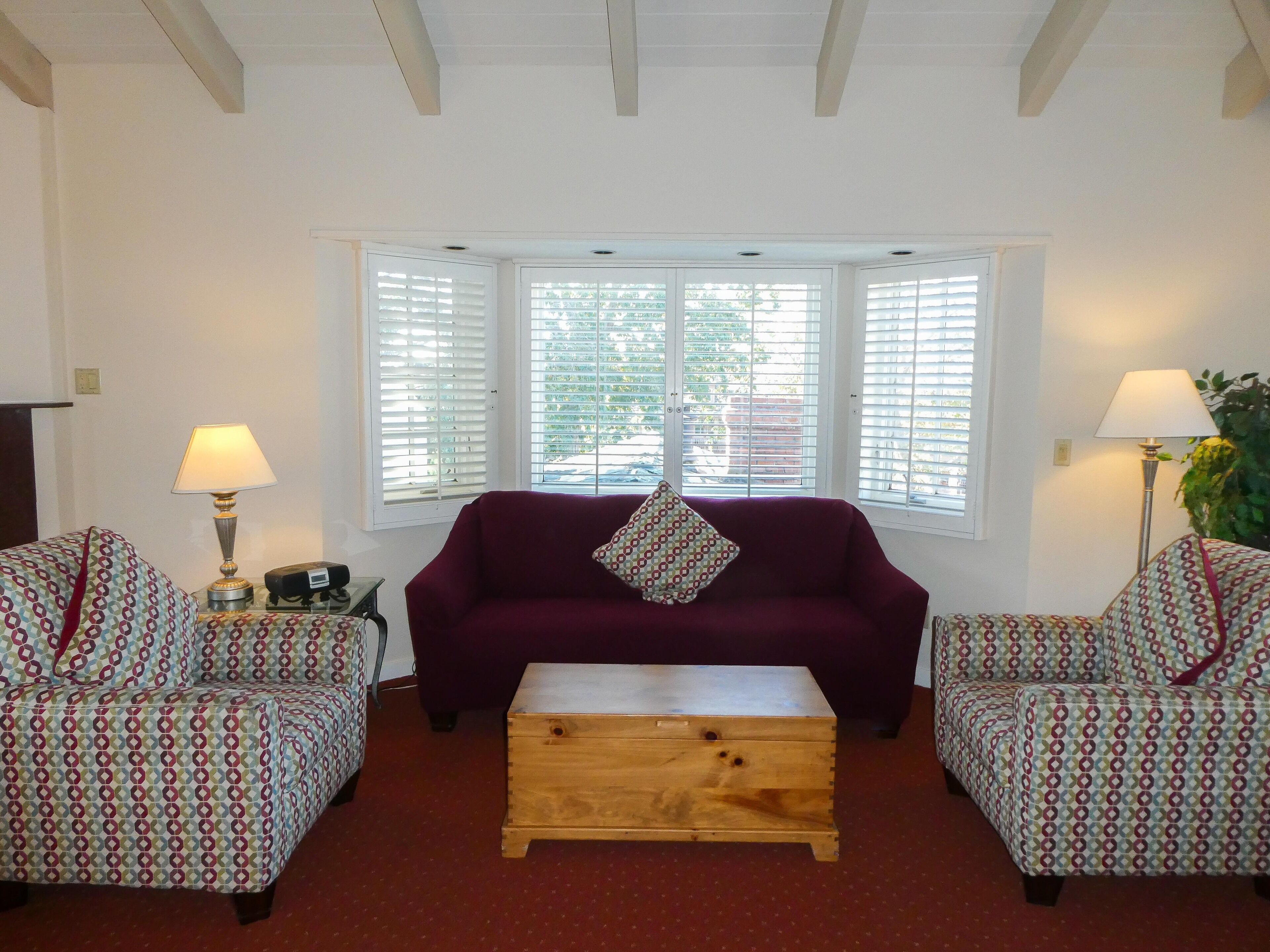 Una sala de estar con un sofá morado, dos sillones estampados, una mesa de centro de madera y un ventanal con contraventanas blancas.