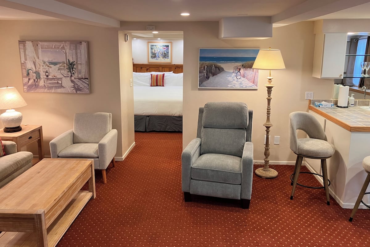 A hotel room with a bed, two armchairs, a wooden coffee table, a floor lamp, a desk with a chair, and artwork on the walls.