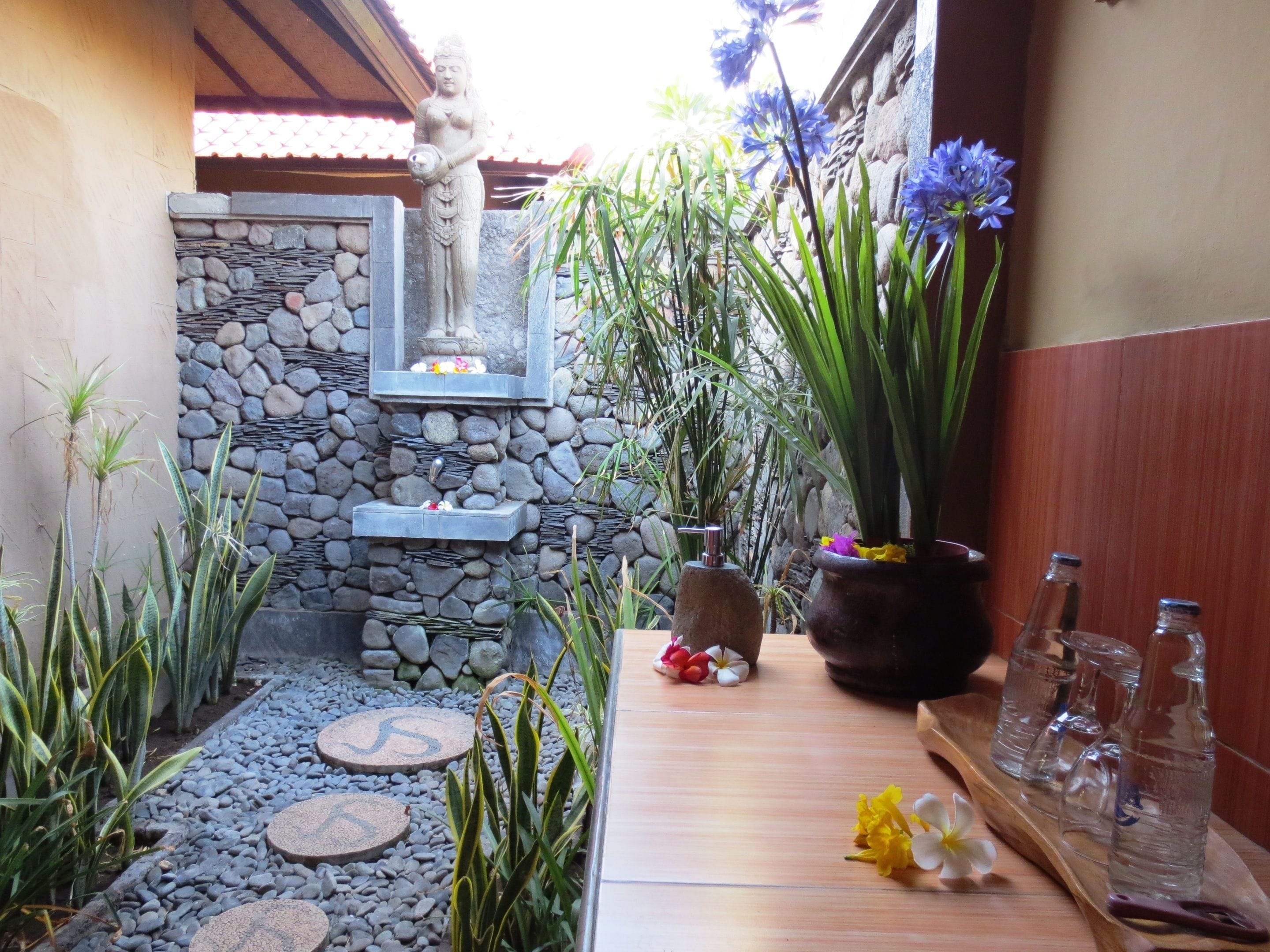 Un espacio exterior tranquilo con un muro de piedra, una fuente de agua pequeña y una mesa de madera con botellas de vidrio y flores.