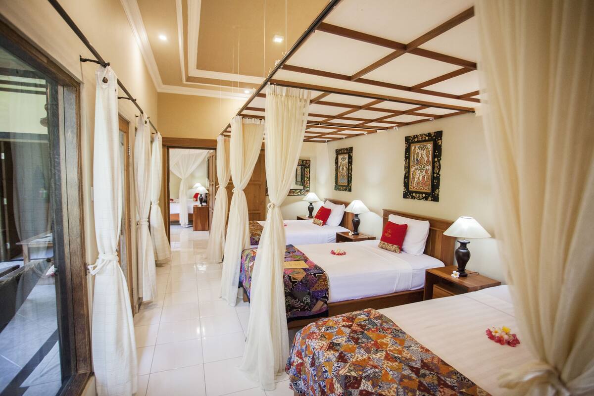 A hotel room with four beds, each with a different patterned bedspread. There are curtains on the windows, and the room has a tiled floor.