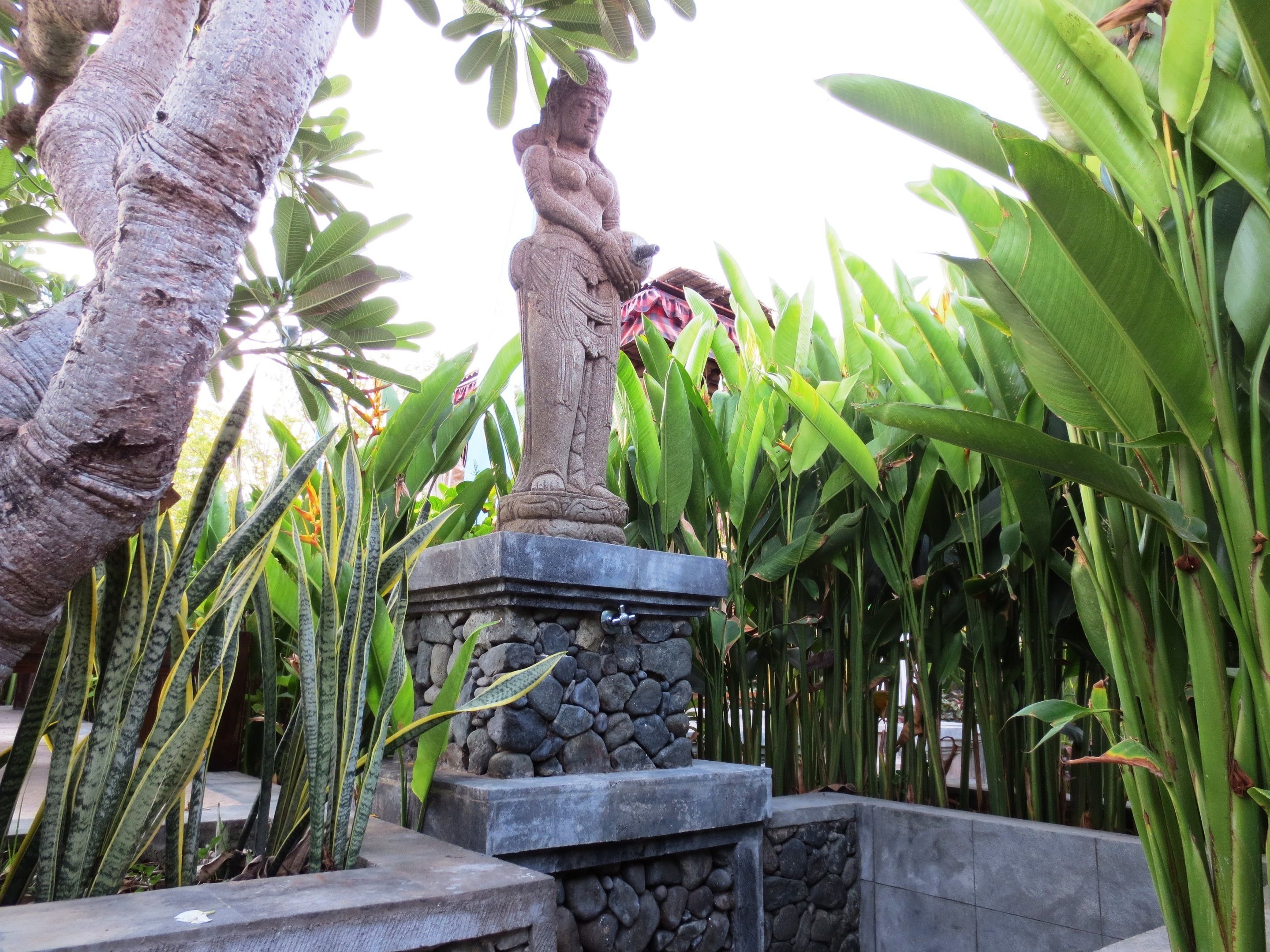 Una estatua de piedra con una figura sentada en un pedestal, rodeada de follaje tropical.