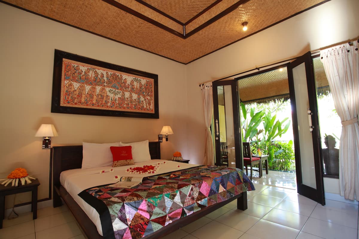 A bedroom with a large bed, two bedside tables, a nightstand, a large framed picture, and a view of a garden through an open door.