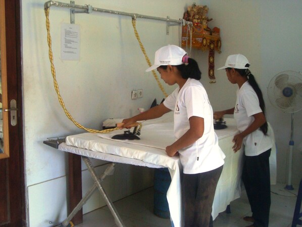 Dos personas planchando ropa en una habitación con una mesa blanca, un ventilador y un adorno en la pared.