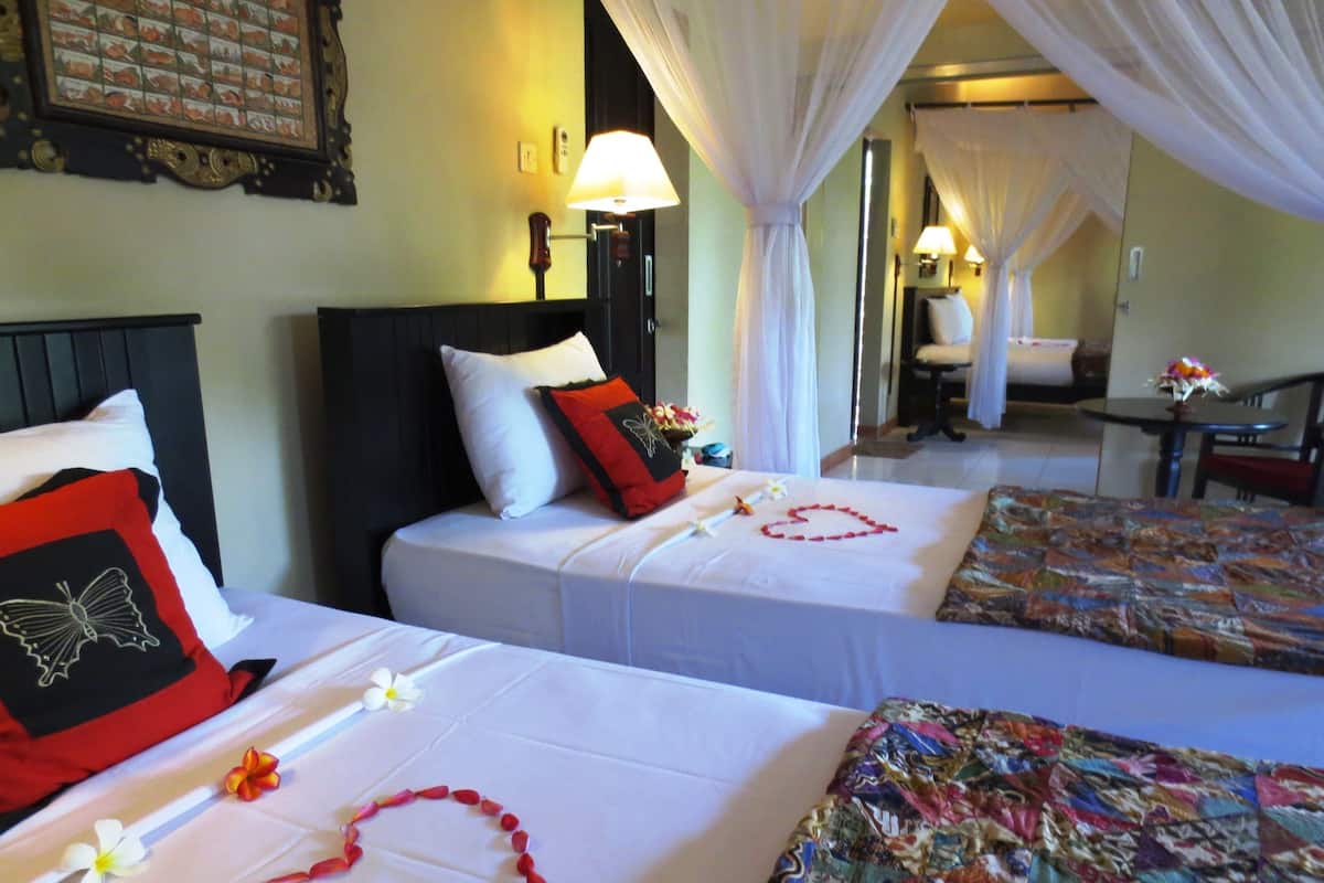 A hotel room with two beds, each with a white bedspread and a red pillow. There are decorative flowers and a heart-shaped arrangement on the beds. The room has a canopy bed and a small table with a lamp.