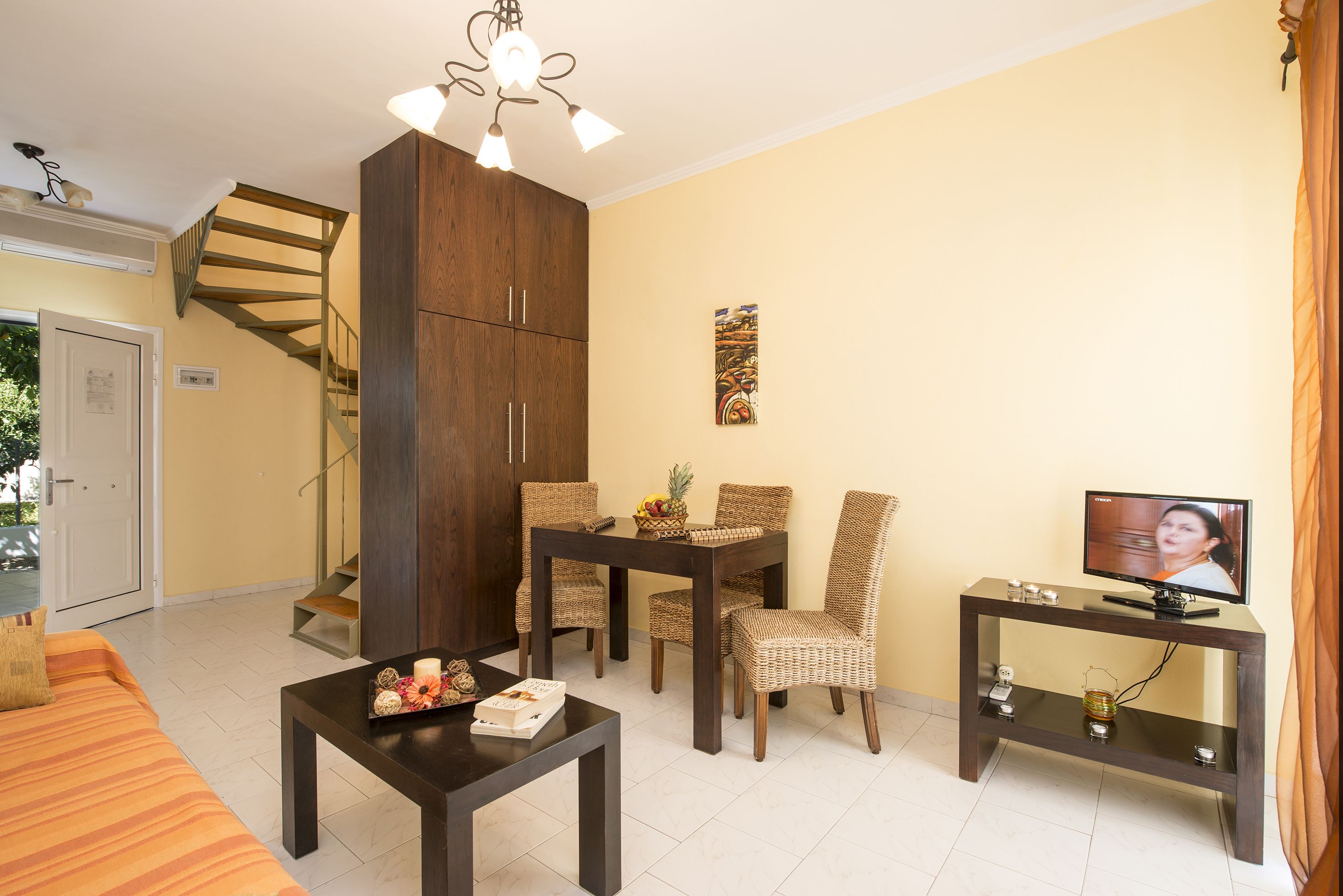 Un salon comprenant un canapé, une table à manger avec des chaises, un téléviseur posé sur un meuble, un escalier et une grande armoire en bois.