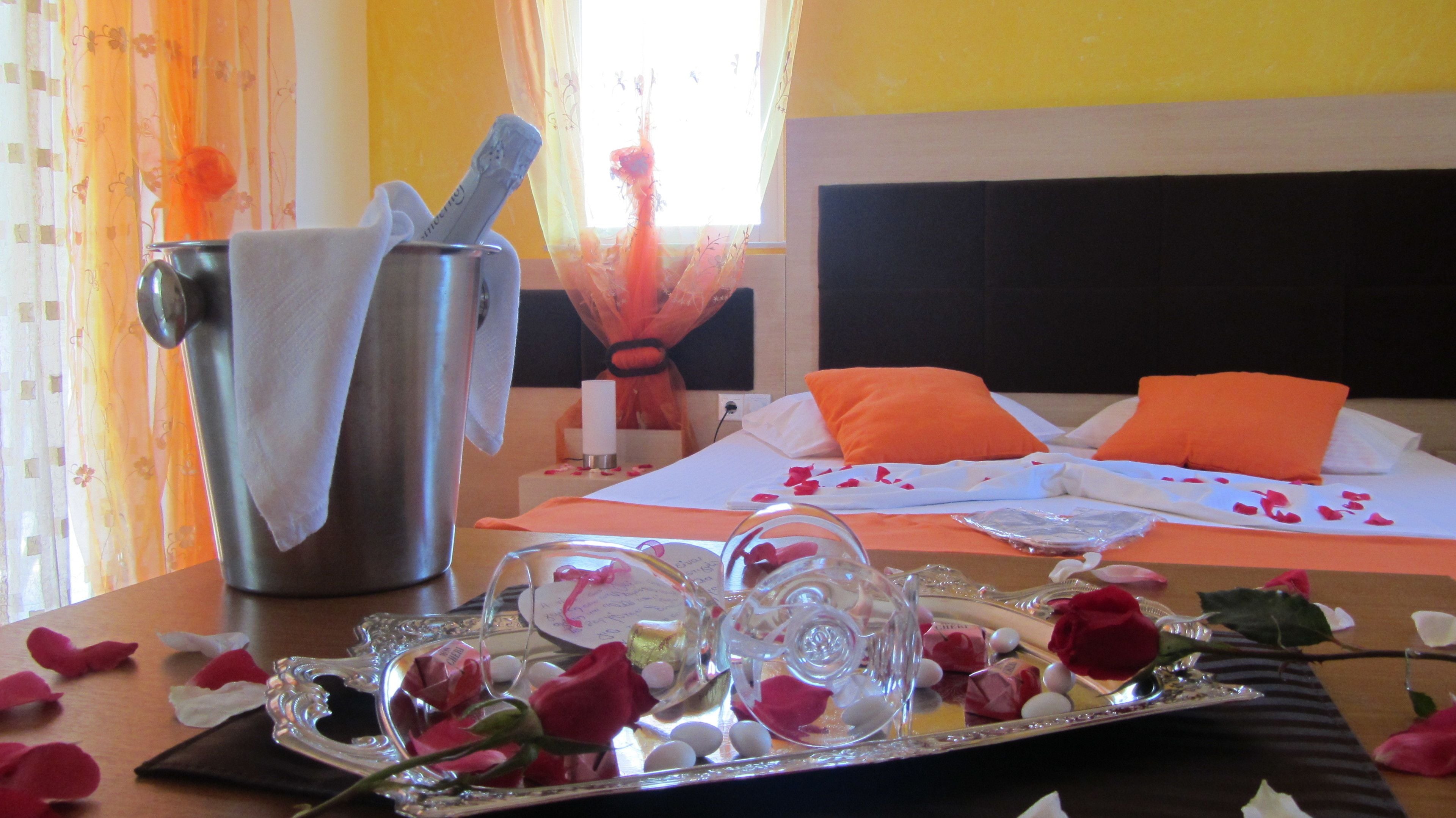 A hotel room with a bed, orange pillows, a bedside table with a glass vase of flowers, a tray with rose petals, and a metal container with a towel.