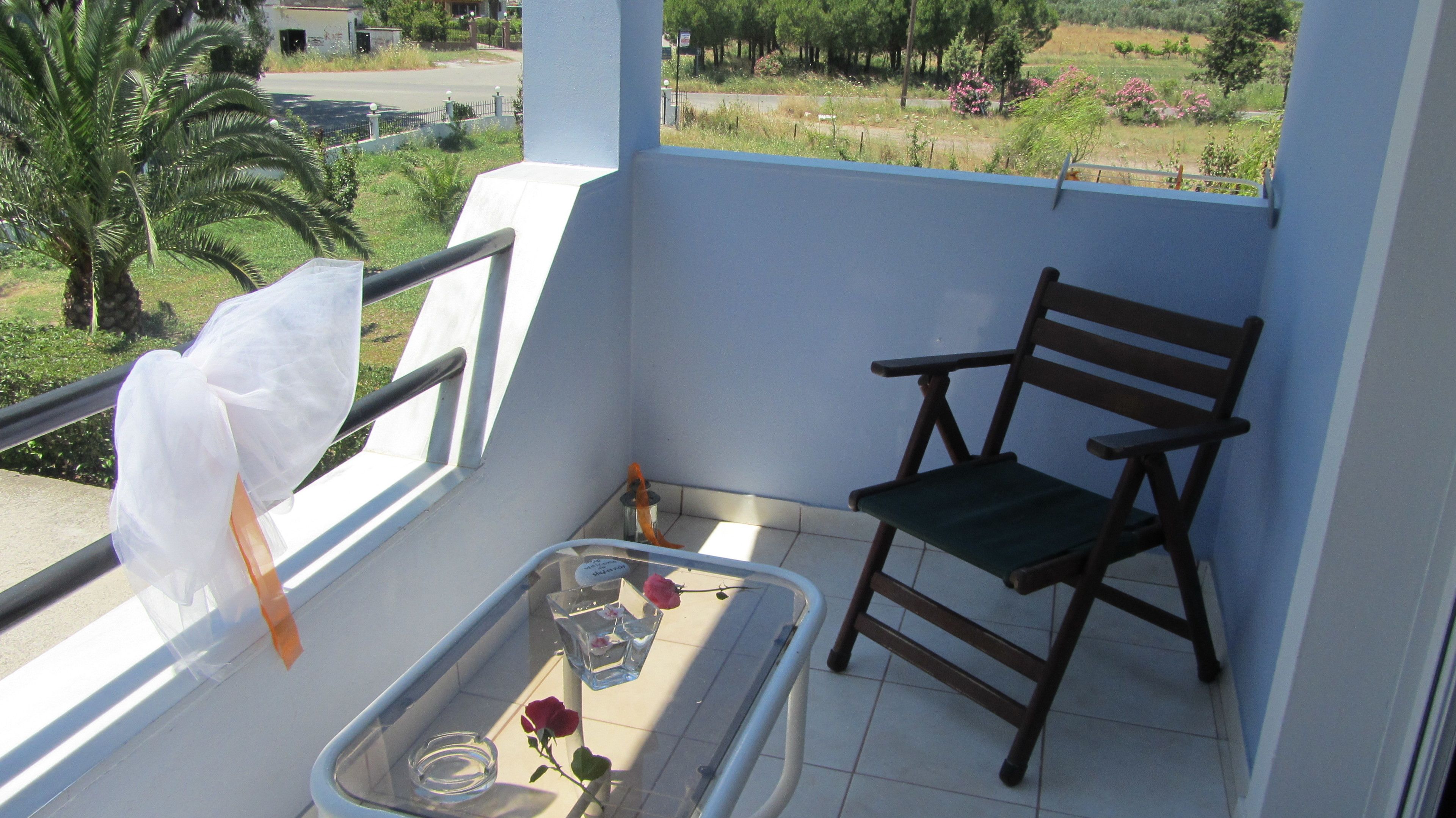 Un balcón con una silla, una mesita con un florero de vidrio y una rosa, y vista a un paisaje verde con árboles y una carretera.