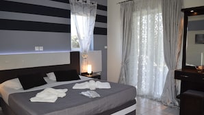 A modern hotel room with a large bed, a bedside table with a lamp, a dresser, and a window with curtains. - Shine Studios (Lesvos)
