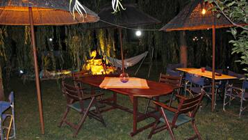 Een terras met houten tafels en stoelen, omgeven door rieten parasols, en een hangmat op de achtergrond.