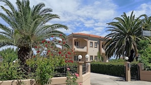 A two-story building with a red-tiled roof, surrounded by palm trees and a well-maintained garden with red flowers. - Shine Studios (Lesvos)