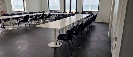 Une salle de conférence avec une longue table et des chaises, de grandes fenêtres offrant une vue dégagée et un sol carrelé.
