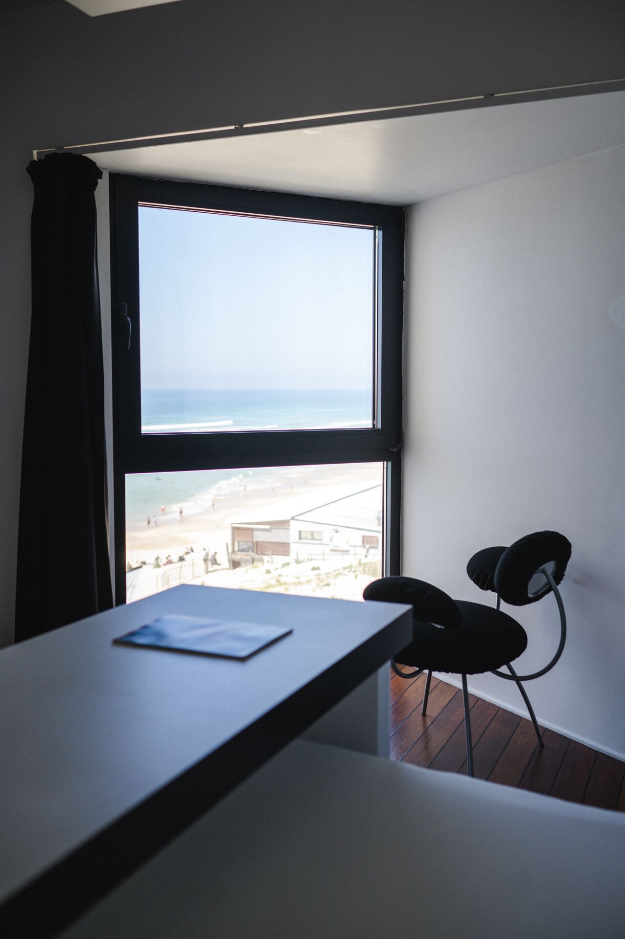Una habitación con un ventanal grande que da a una playa, un escritorio con una silla y una cortina.