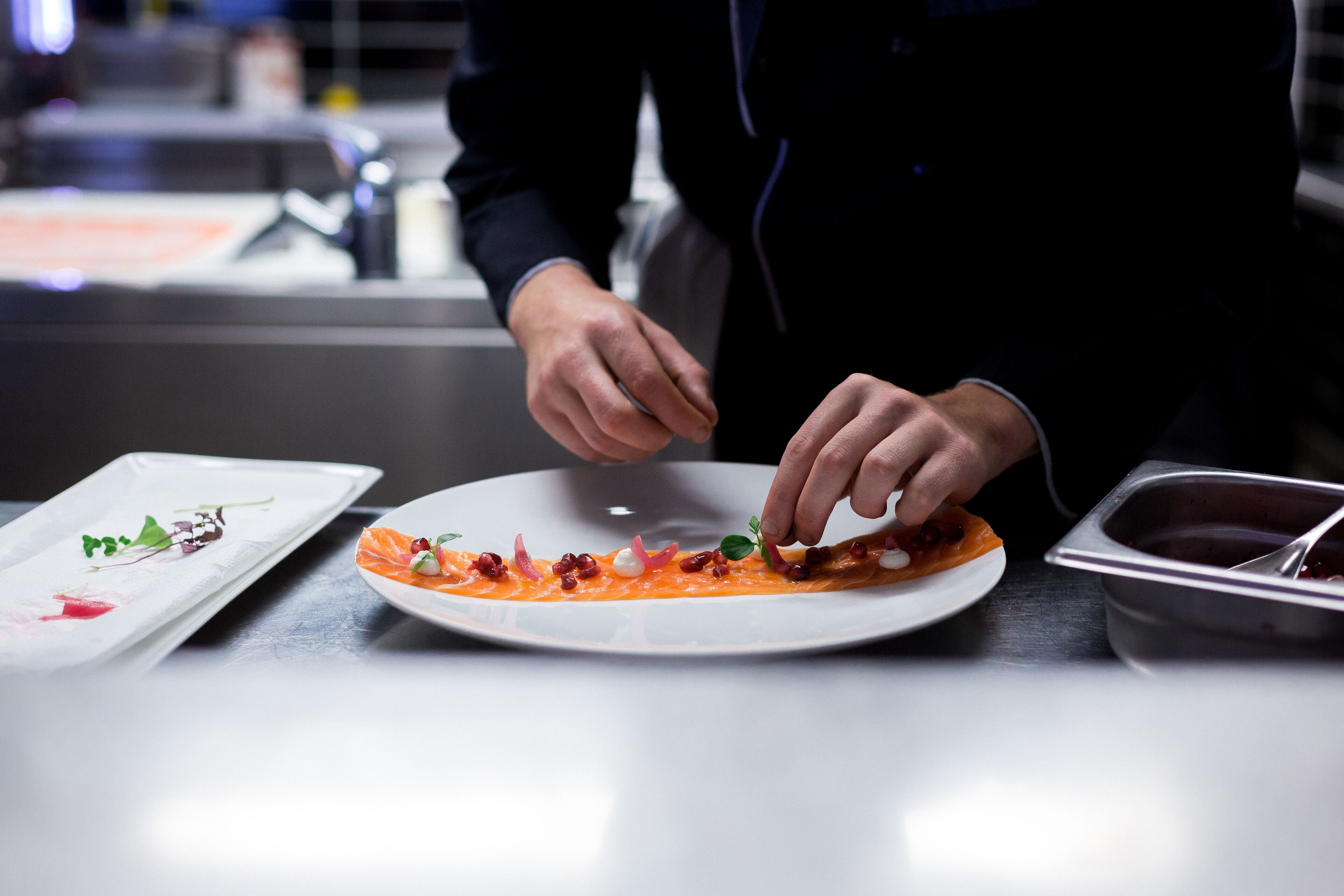 Un chef preparando un plato con una fuente blanca y un recipiente metálico sobre una encimera.