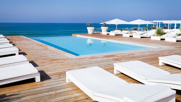 Un espace aménagé au bord de la piscine, avec des chaises longues blanches et des parasols, une terrasse en bois et une vue sur l’océan.
