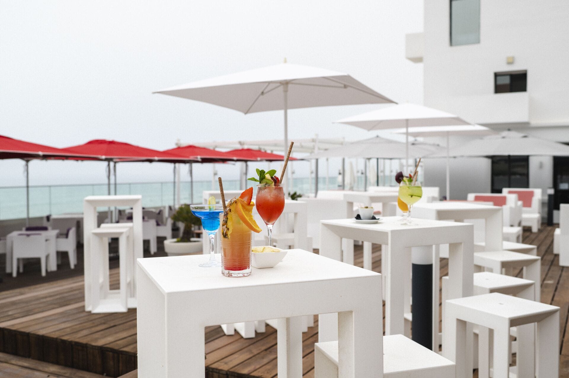 Eine Dachterrasse mit weißen Tischen und roten Sonnenschirmen mit Blick aufs Meer.