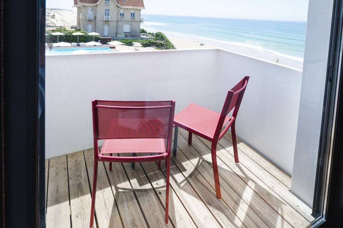 Un balcon doté d’une terrasse en bois, de deux chaises rouges et offrant une vue sur une plage et un bâtiment situé au loin.