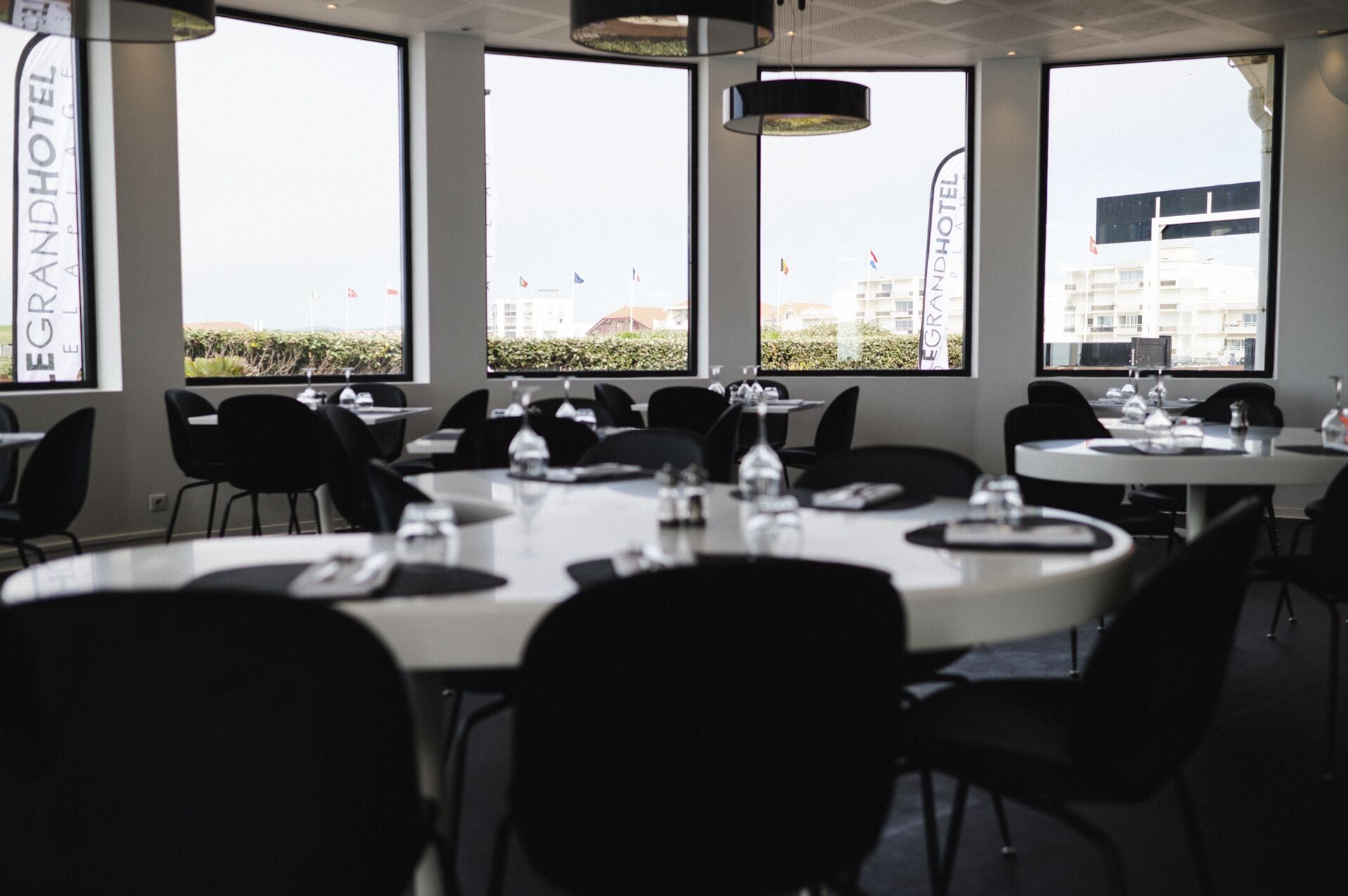 A dining area with round tables and black chairs, large windows with views of buildings, and a sign reading "EGRANDHOTEL".