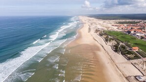 Une vue aérienne d’une ville côtière avec une vaste plage de sable, une mer bleue et limpide, et un mélange de bâtiments résidentiels et commerciaux.