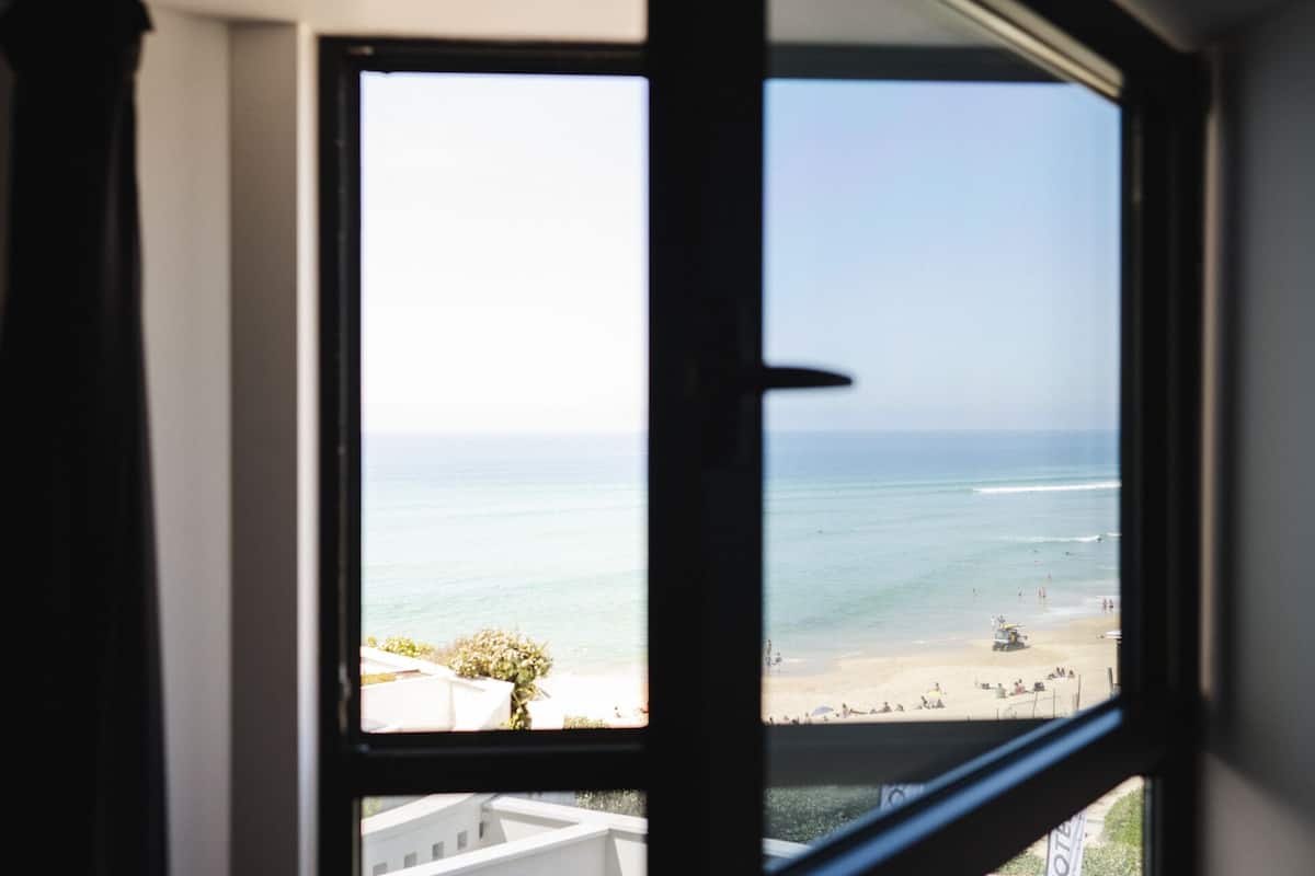Une vue sur une plage, prise à travers une fenêtre, avec l'océan et les personnes se promenant sur la plage en arrière-plan.